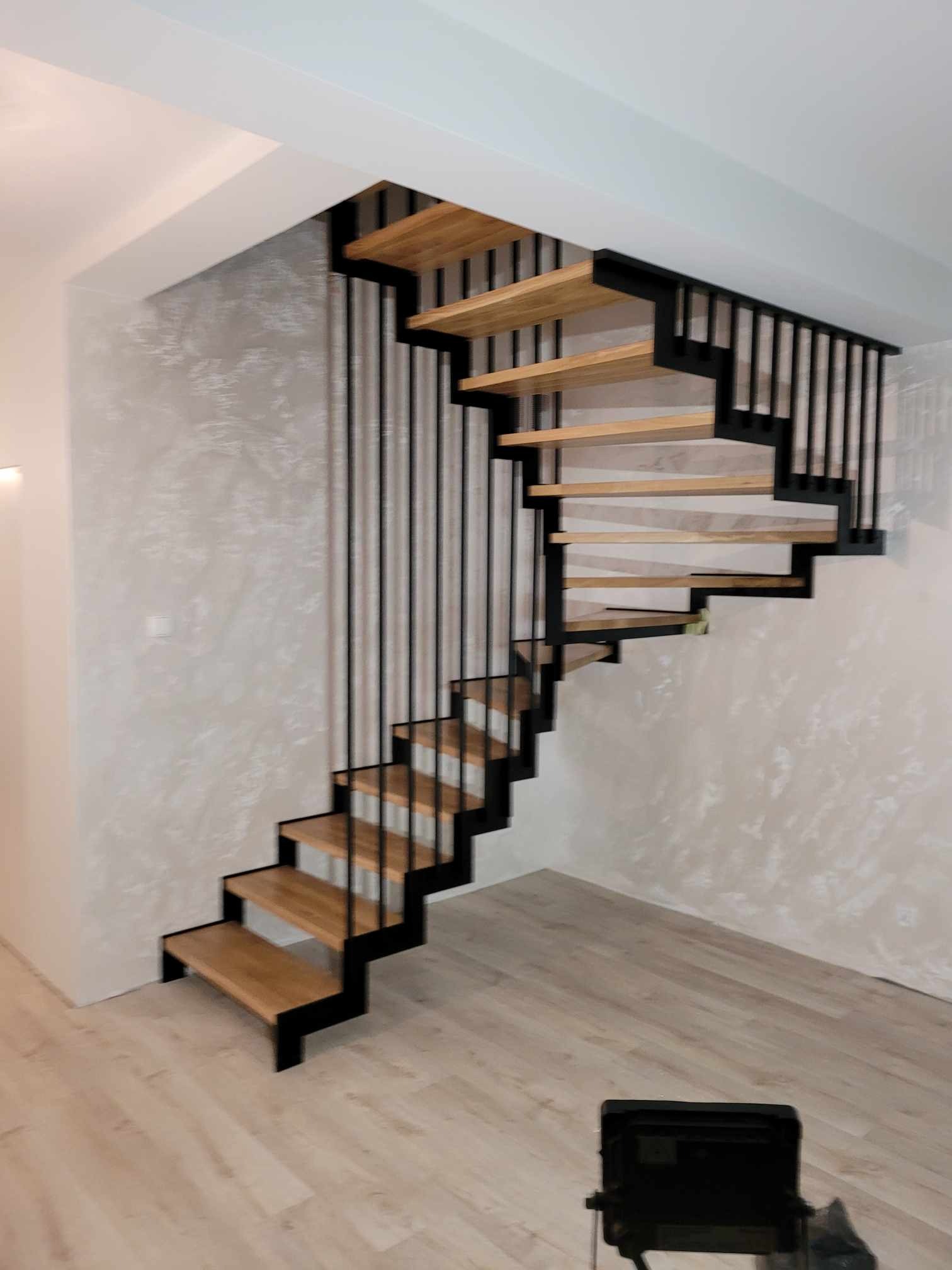 Nowoczesne schody ażurowe z drewnianymi stopniami i czarną metalową konstrukcją w jasnym wnętrzu z dekoracyjną ścianą.