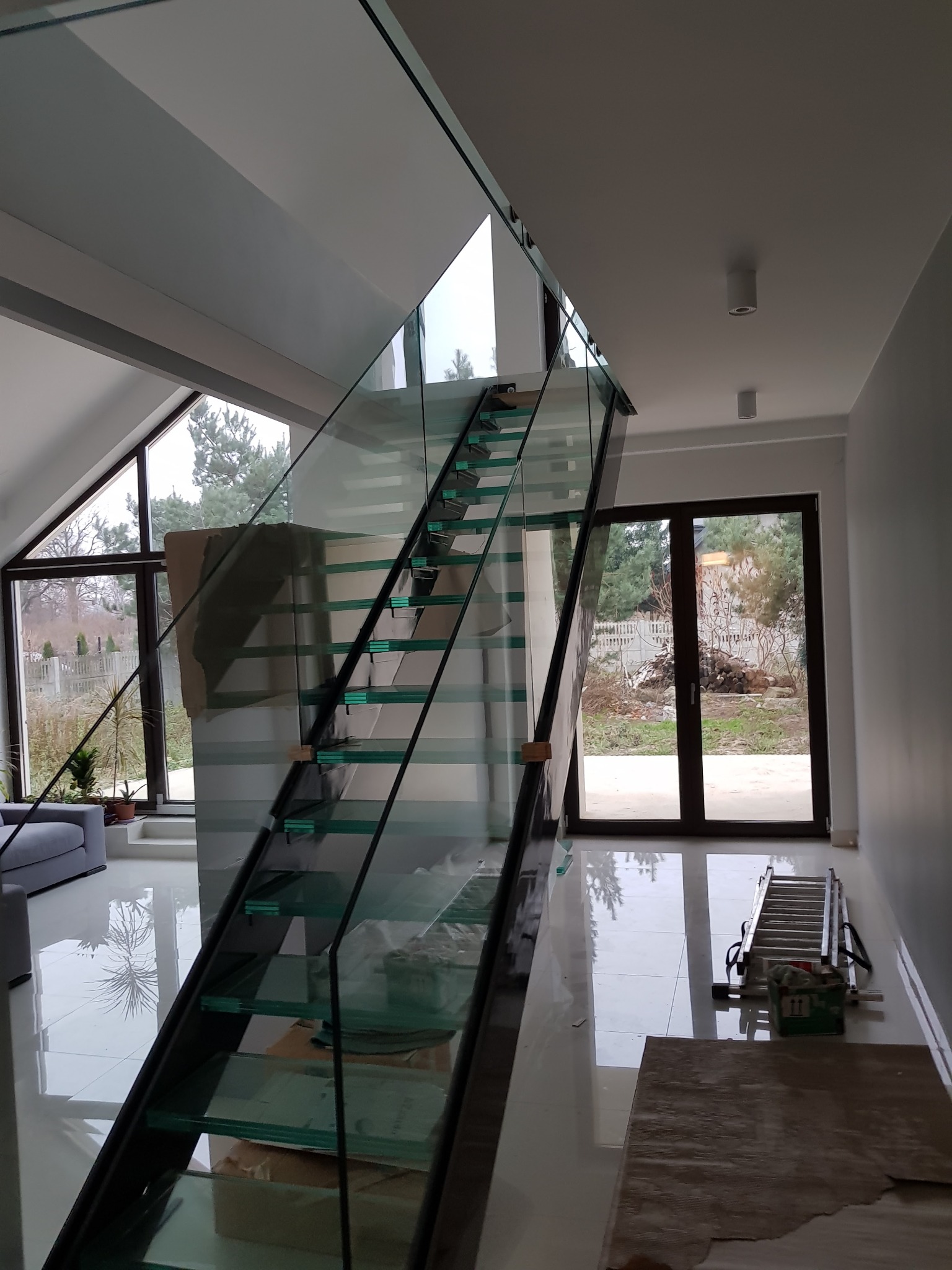 Nowoczesne, szklane schody z metalową konstrukcją w jasnym wnętrzu domu w Maksymilianowie, widoczne z poziomu podłogi, z elementami zabezpieczającymi na szybach i drabiną opartą o ścianę.