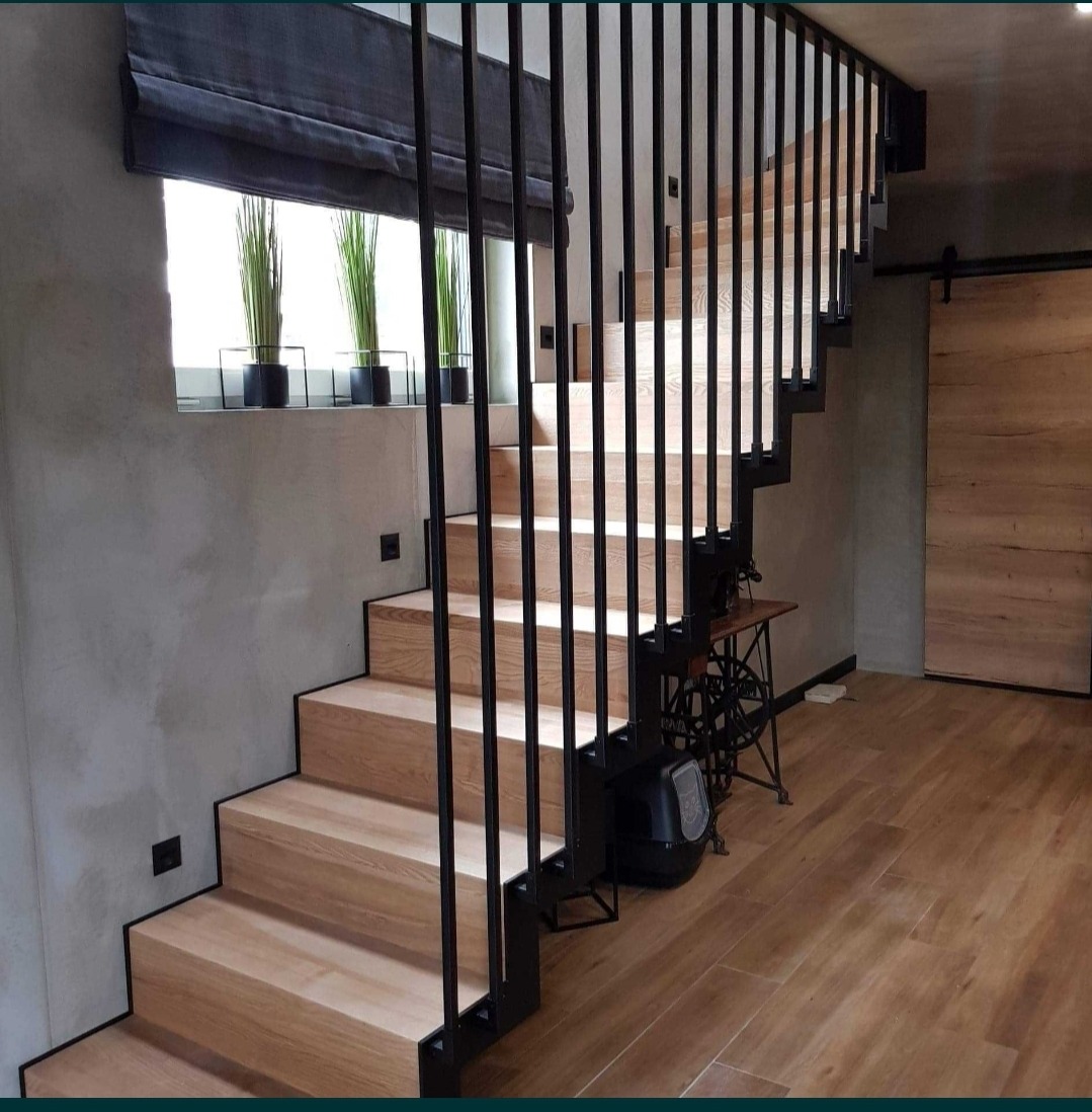 Nowoczesne schody z jasnego drewna z czarną metalową balustradą, widok z dołu na tle szarej ściany i drewnianych drzwi przesuwnych.