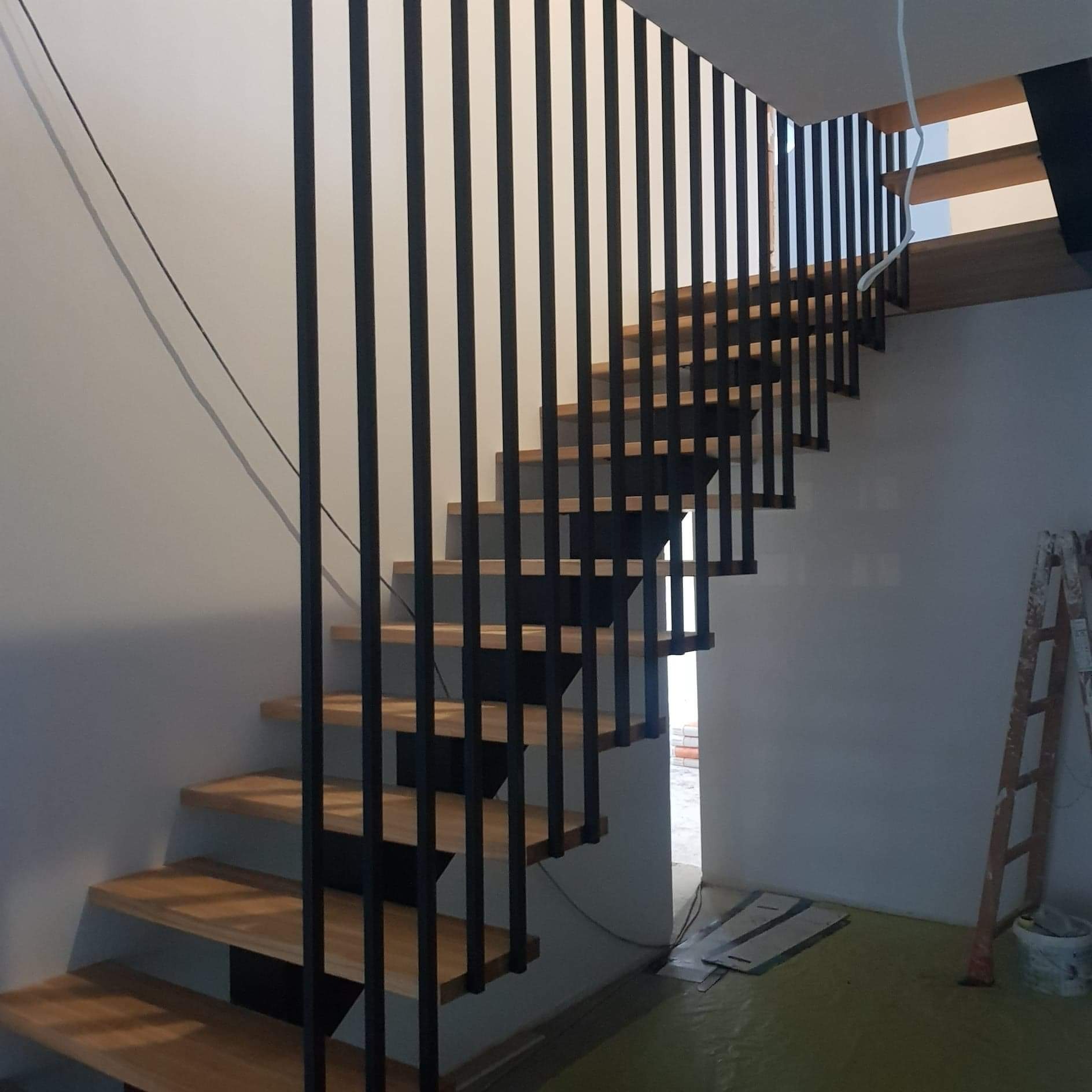 Nowoczesne schody z drewnianymi stopniami i czarną metalową balustradą z pionowych prętów, widoczne elementy wykończeniowe i drabina malarska w tle.
