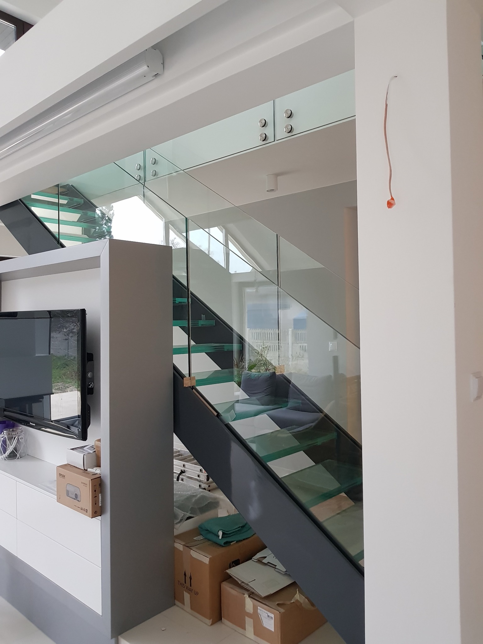 Szklane schody z metalową konstrukcją w nowoczesnym wnętrzu, widoczne niedokończone prace wykończeniowe, w tle salon z oknami.