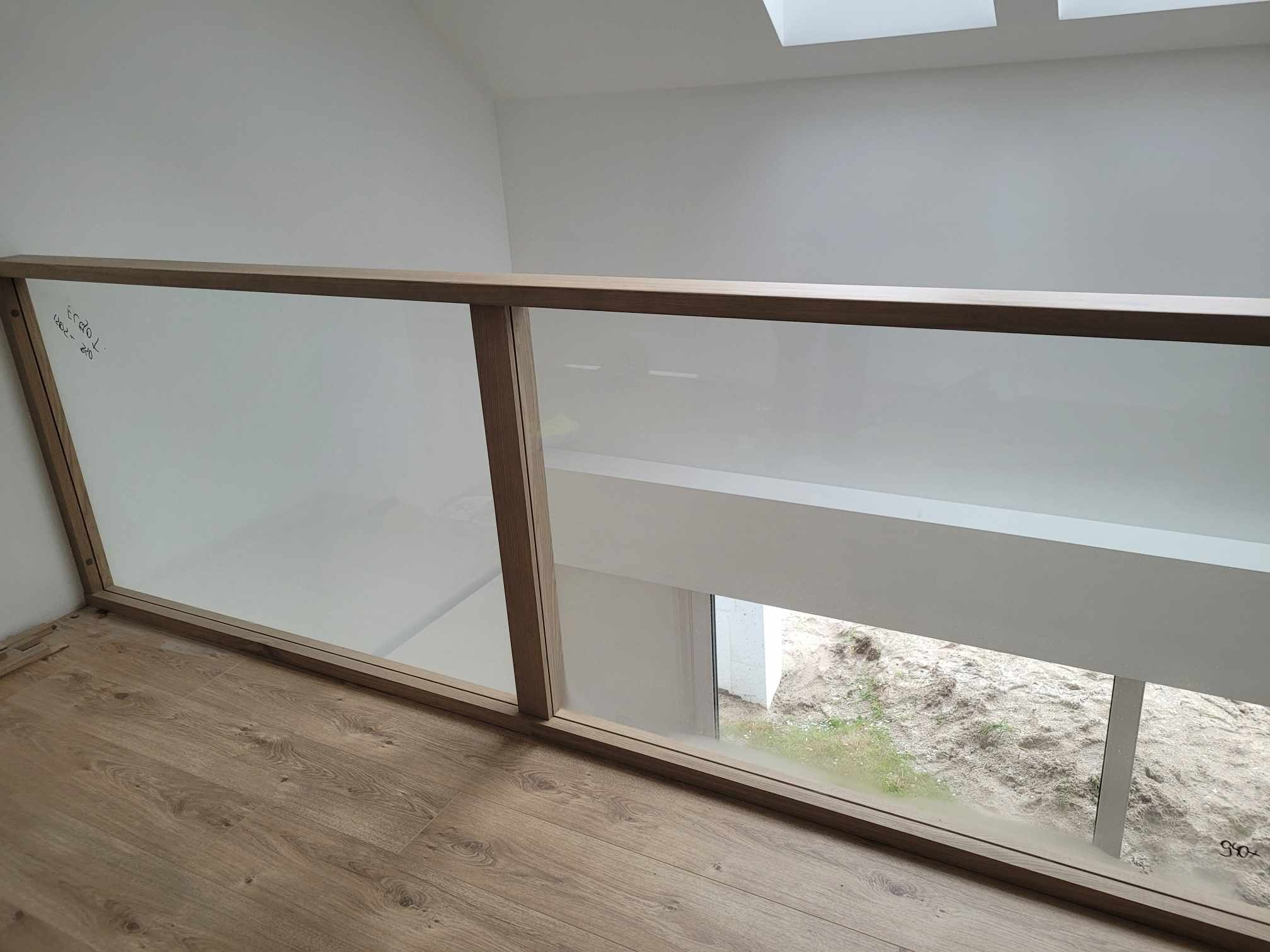 Szklana balustrada z drewnianą ramą na antresoli, widok na zewnątrz, jasne drewno, minimalistyczny design, widoczne notatki na szkle.