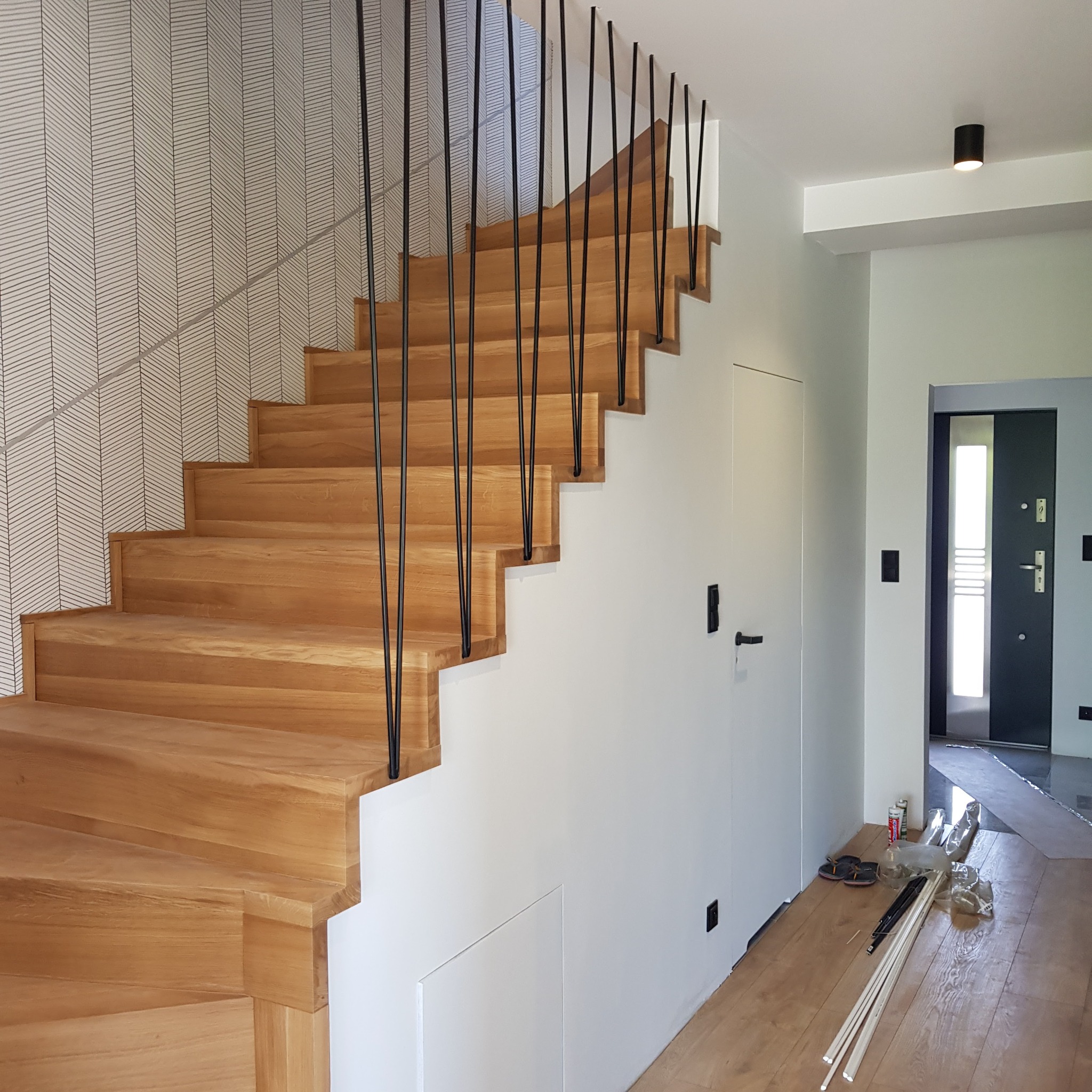 Nowoczesne schody z jasnego drewna z czarnymi, pionowymi balustradami, przylegające do ściany z geometryczną tapetą, widoczne materiały wykończeniowe na podłodze.