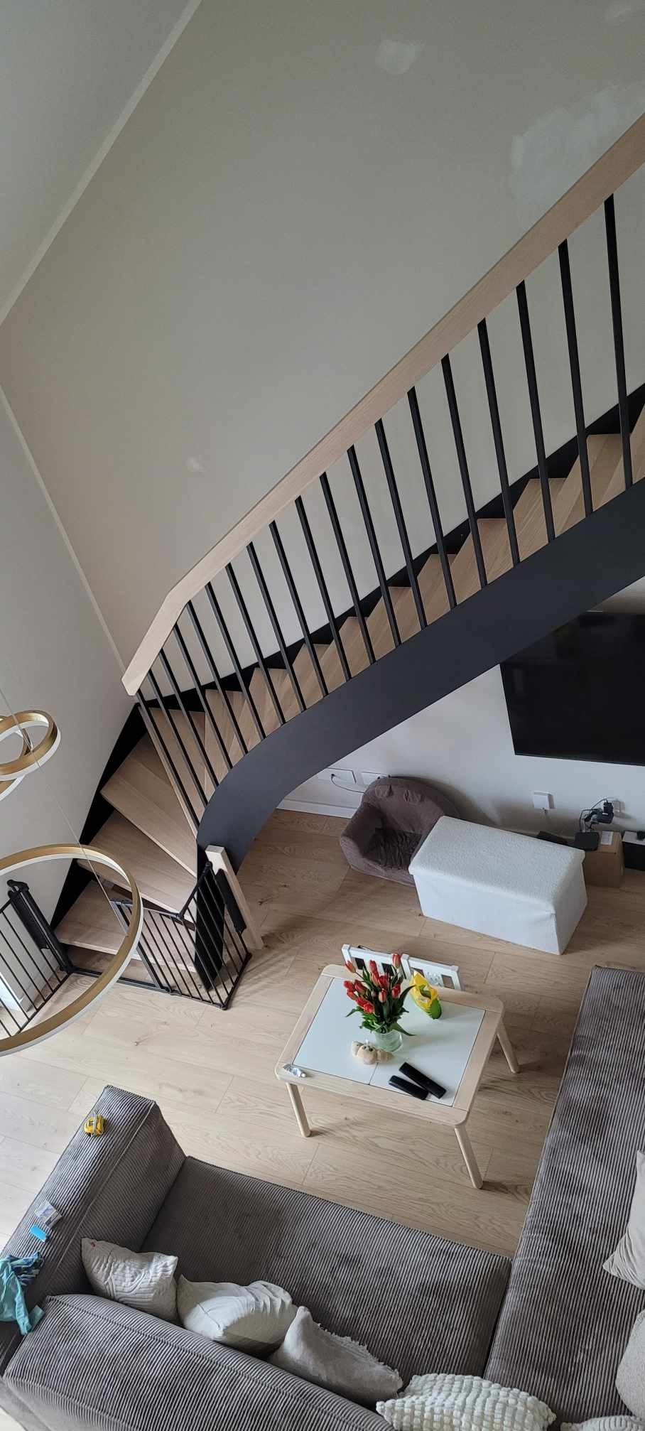 Widok z góry na nowoczesne schody z drewnianymi stopniami i czarną metalową balustradą, umieszczone w jasnym wnętrzu z drewnianą podłogą, widoczne także fragmenty salonu z sofą, stolikiem kawowym...