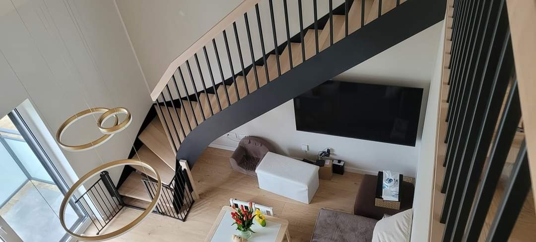 Wnętrze domu z widokiem z góry na drewniane schody z czarną balustradą, nowoczesne lampy wiszące w kształcie okręgów, telewizor na ścianie i meble w salonie.