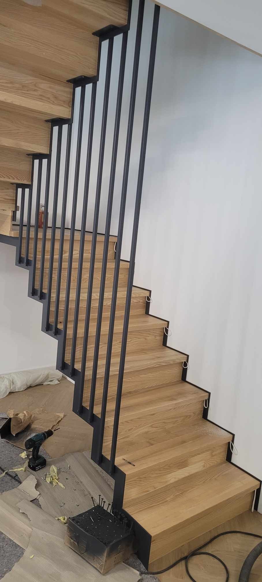 Montaż drewnianych schodów z czarną, metalową balustradą; widoczne narzędzia i materiały budowlane na podłodze.
