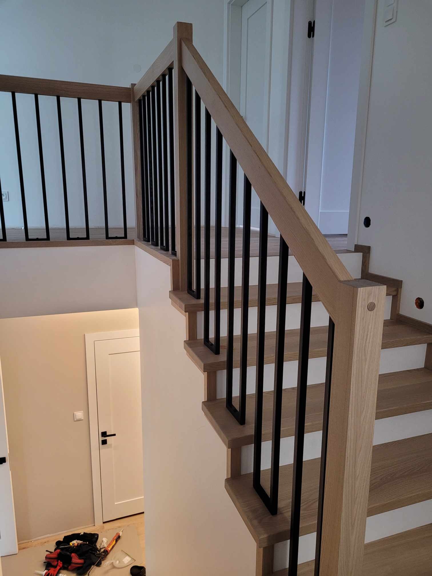Nowoczesne schody z drewnianymi stopniami i balustradą z czarnymi, pionowymi elementami, widoczne z boku, z narzędziami budowlanymi w tle.