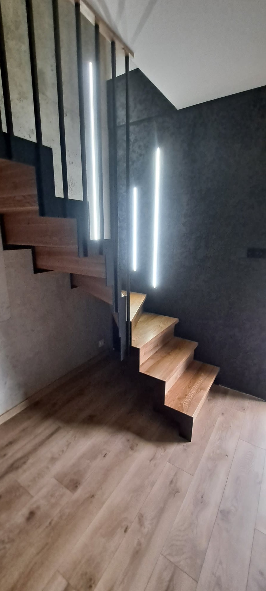 Nowoczesne schody z drewnianymi stopniami i metalową balustradą, oświetlone pionowymi lampami LED na ciemnej ścianie.