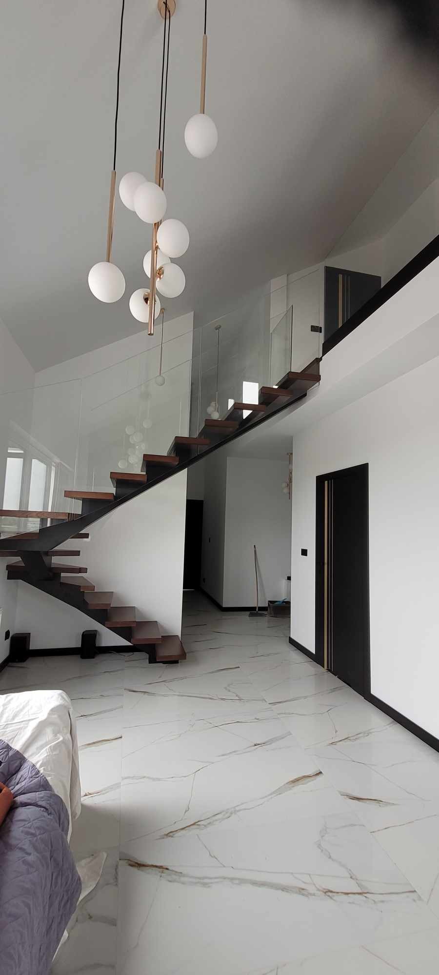 Nowoczesne schody z drewnianymi stopniami i szklanymi balustradami w przestronnym wnętrzu z antresolą, oświetlone designerską lampą wiszącą.