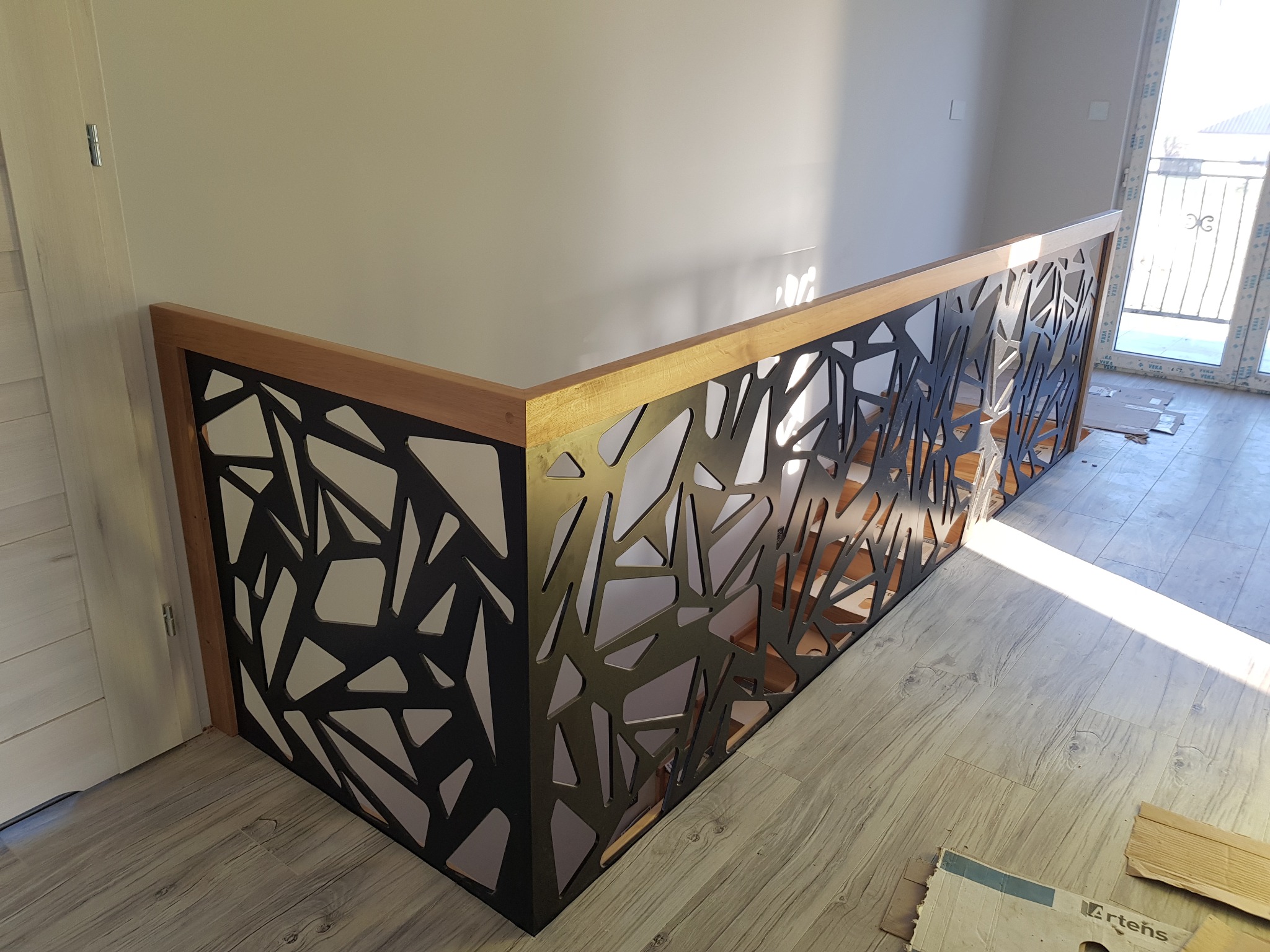 Nowoczesna balustrada z czarnego metalu o geometrycznym wzorze, osadzona na drewnianej ramie, oświetlona naturalnym światłem w jasnym wnętrzu.