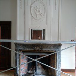 Bud In Nenad Jandric - Klasyczny kominek z dekoracyjnym obramowaniem i płaskorzeźbą na ścianie, w trakcie prac renowacyjnych, widoczne rusztowanie i taśma zabezpieczająca na podłodze.