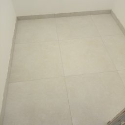 Bud In Nenad Jandric - Ułożone jasne płytki podłogowe w łazience, widoczne fragmenty rur na ścianie i listwy przypodłogowe.