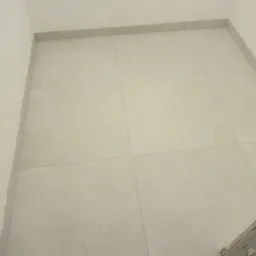 Ułożone jasne płytki podłogowe w łazience, widoczne fragmenty rur na ścianie i listwy przypodłogowe.