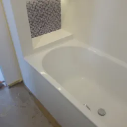 Biała wanna po renowacji, z mozaiką w strefie prysznicowej i półką, widoczne ślady prac wykończeniowych na podłodze.