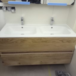 Bud In Nenad Jandric - Nowoczesna umywalka podwójna z drewnianą szafką i chromowaną armaturą, widoczne niedokończone prace instalacyjne i odbicie osoby robiącej zdjęcie w lustrze.