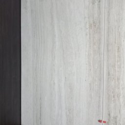 Bud In Nenad Jandric - Ściana wyłożona płytami imitującymi trawertyn, z widocznymi elementami konstrukcji podwieszanej i czerwonymi krzyżykami dystansowymi między płytami.