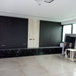 Bud In Nenad Jandric - Wnętrze w trakcie prac wykończeniowych: ściana z panelami w kolorze czarnym i beżowym, marmurowy cokół, jasna podłoga, okna i materiały budowlane w tle.