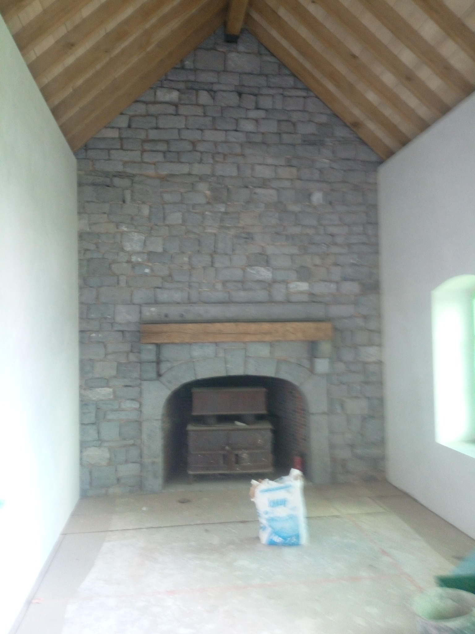 Surowa ściana z kamienia z drewnianym nadprożem nad wnęką kominkową w trakcie prac wykończeniowych, widoczny worek z zaprawą i surowy drewniany strop.