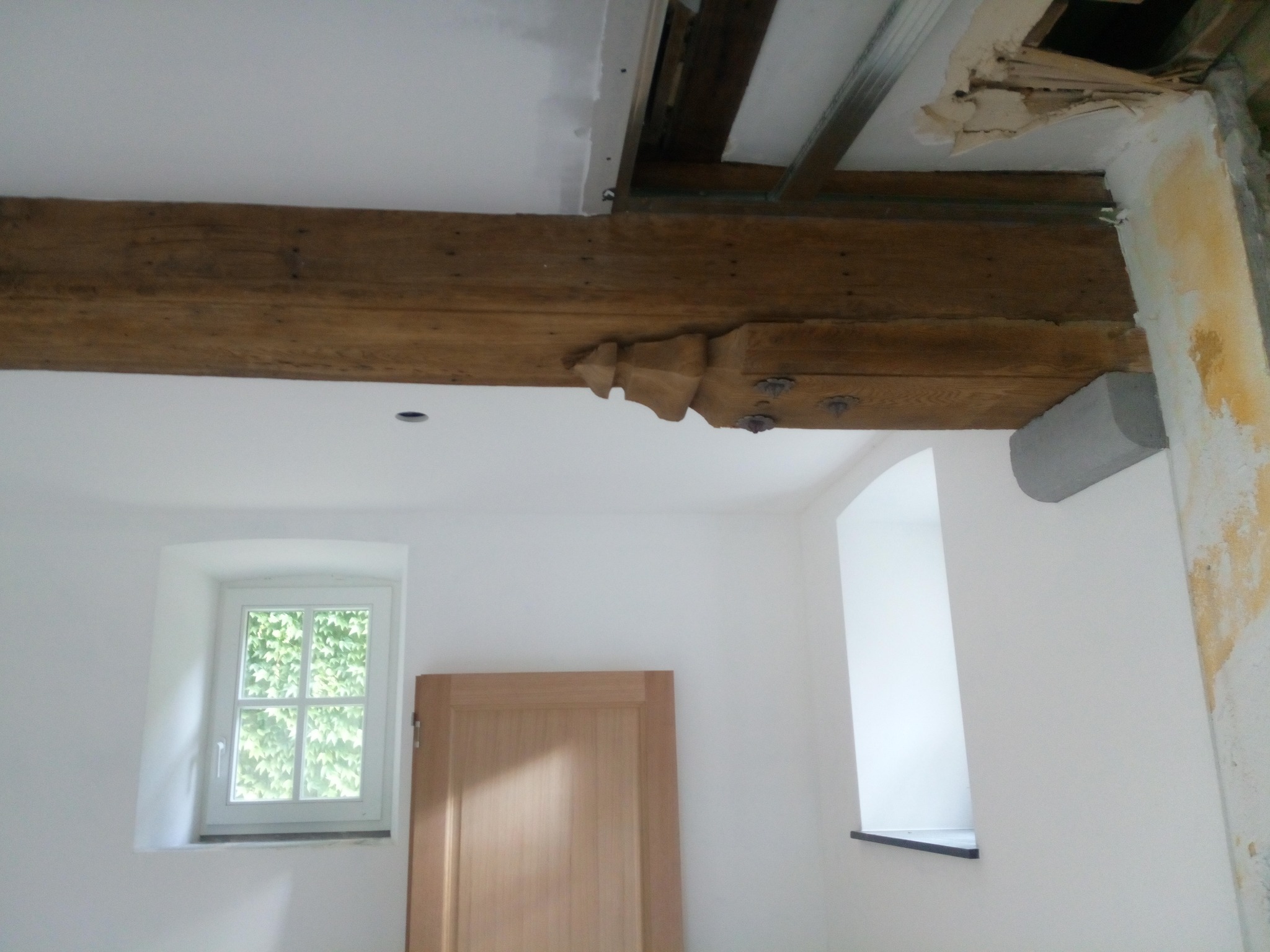 Wnętrze pokoju w trakcie remontu w Rumi: surowa drewniana belka stropowa z dekoracyjnym wykończeniem, widoczne ślady po remoncie na ścianie, przygotowanie do malowania sufitu.
