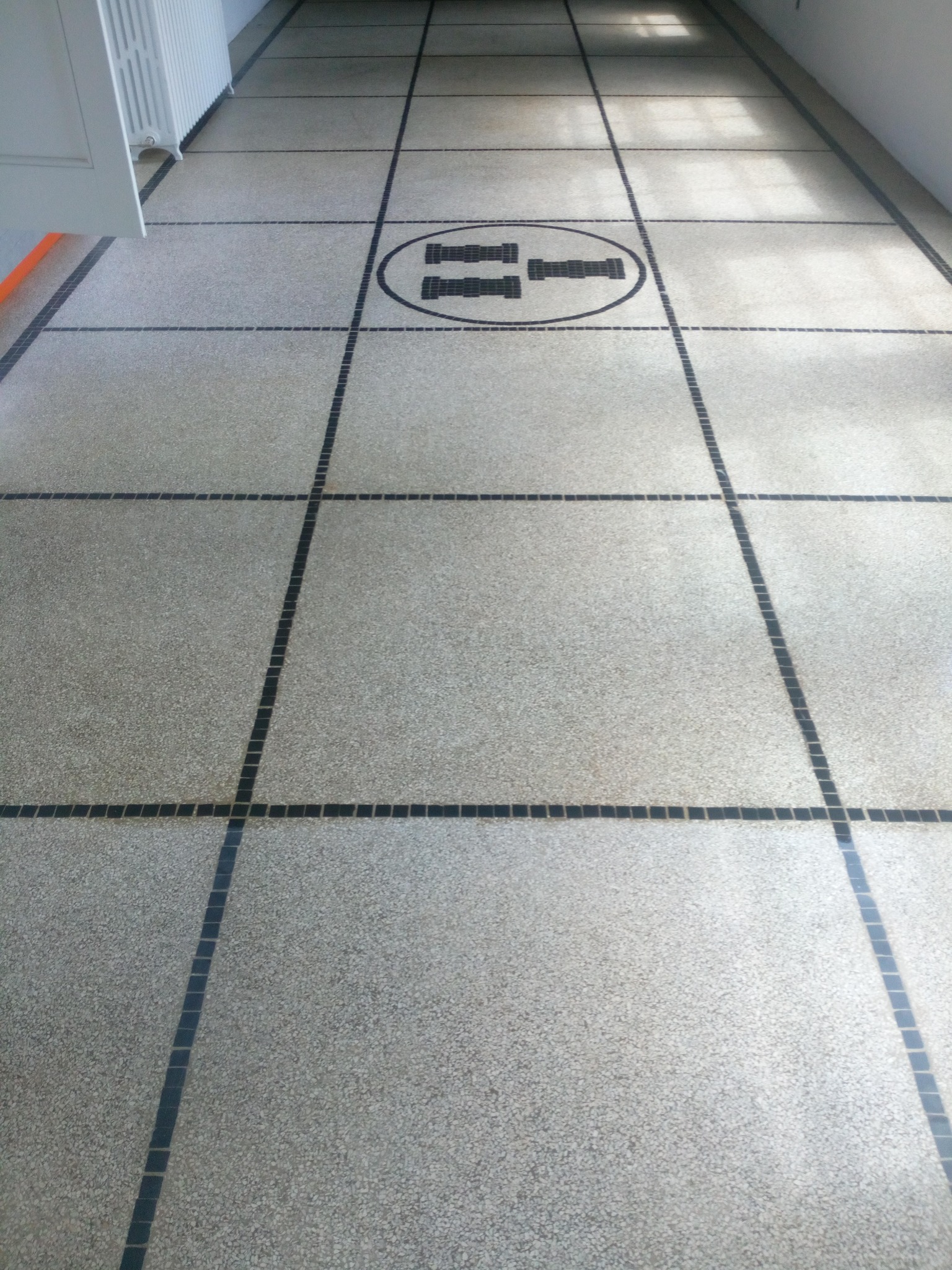 Szara, lastrykowa posadzka z ciemnymi fugami tworzącymi kwadraty i prostokąty, z centralnie umieszczonym okrągłym wzorem z ciemnych elementów.