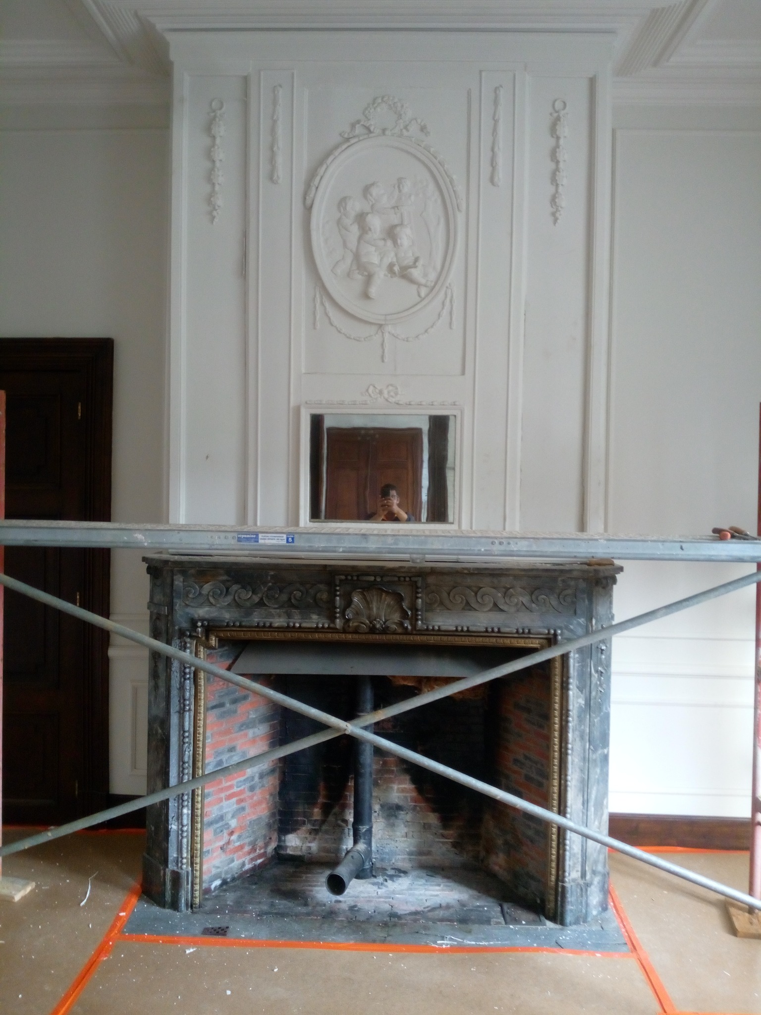 Klasyczny kominek z dekoracyjnym obramowaniem i płaskorzeźbą na ścianie, w trakcie prac renowacyjnych, widoczne rusztowanie i taśma zabezpieczająca na podłodze.
