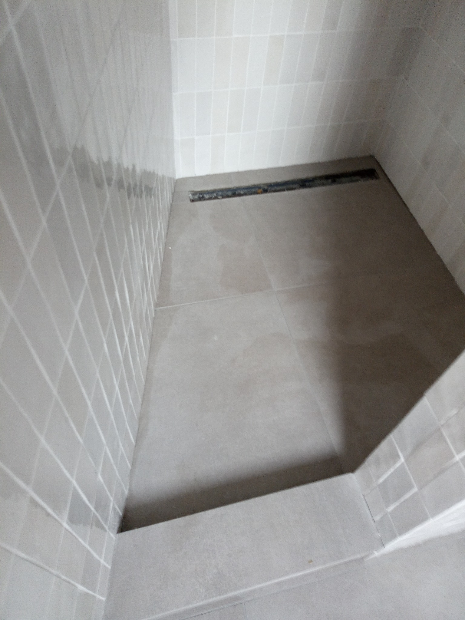 Nowoczesny odpływ liniowy w kabinie prysznicowej wyłożonej jasnymi, kwadratowymi płytkami ceramicznymi na ścianach i podłodze.