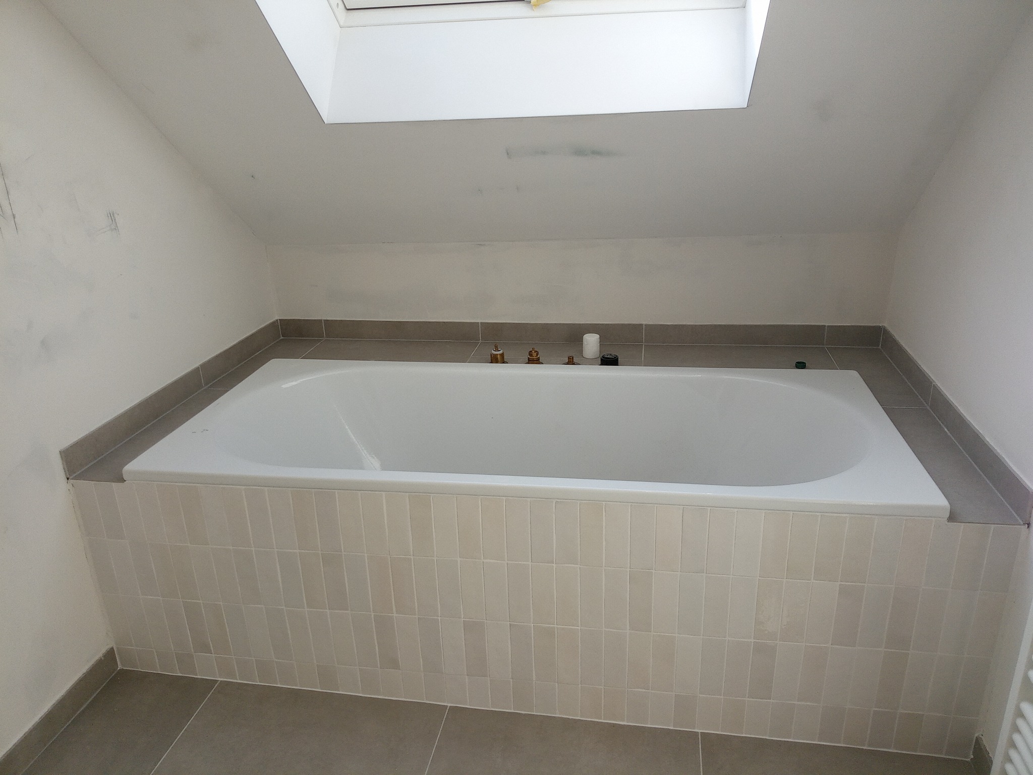 Wanna obudowana płytkami w jasnej łazience na poddaszu ze świetlikiem, widoczne niedoskonałości na ścianie przed malowaniem.