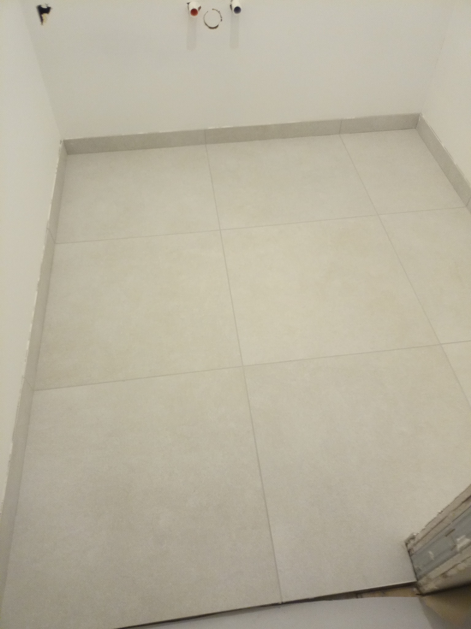 Ułożone jasne płytki podłogowe w łazience, widoczne fragmenty rur na ścianie i listwy przypodłogowe.