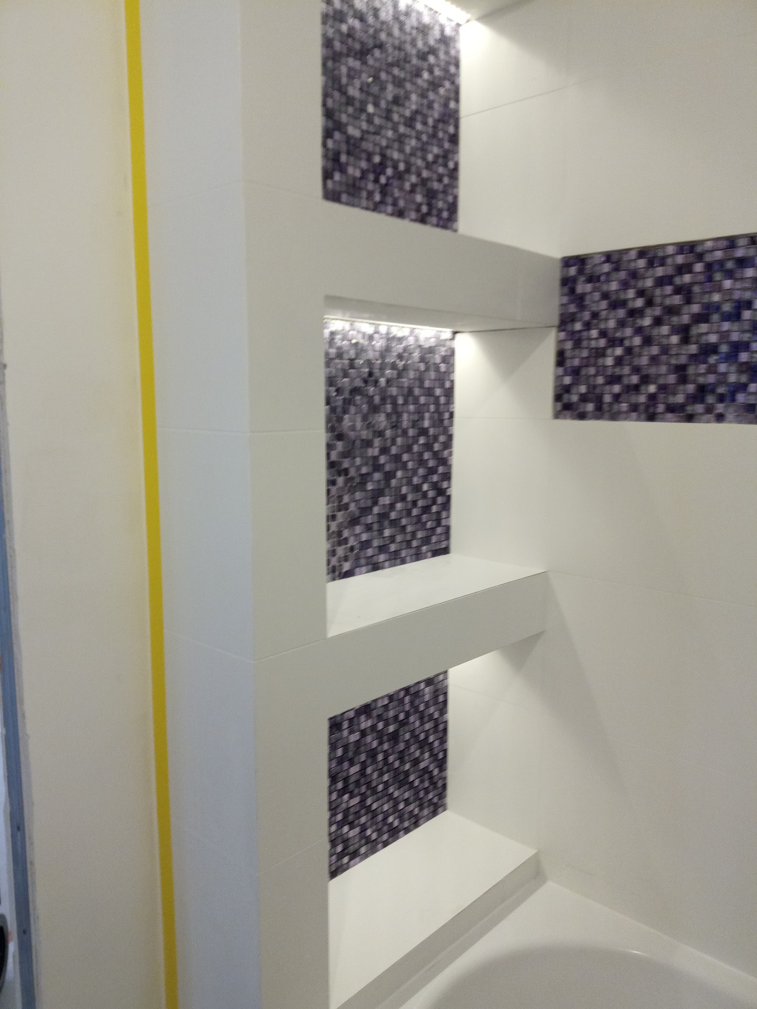 Nowoczesna łazienka z wnękami wyłożonymi mozaiką i podświetleniem LED, widoczne białe płytki i wanna w dolnym rogu kadru.