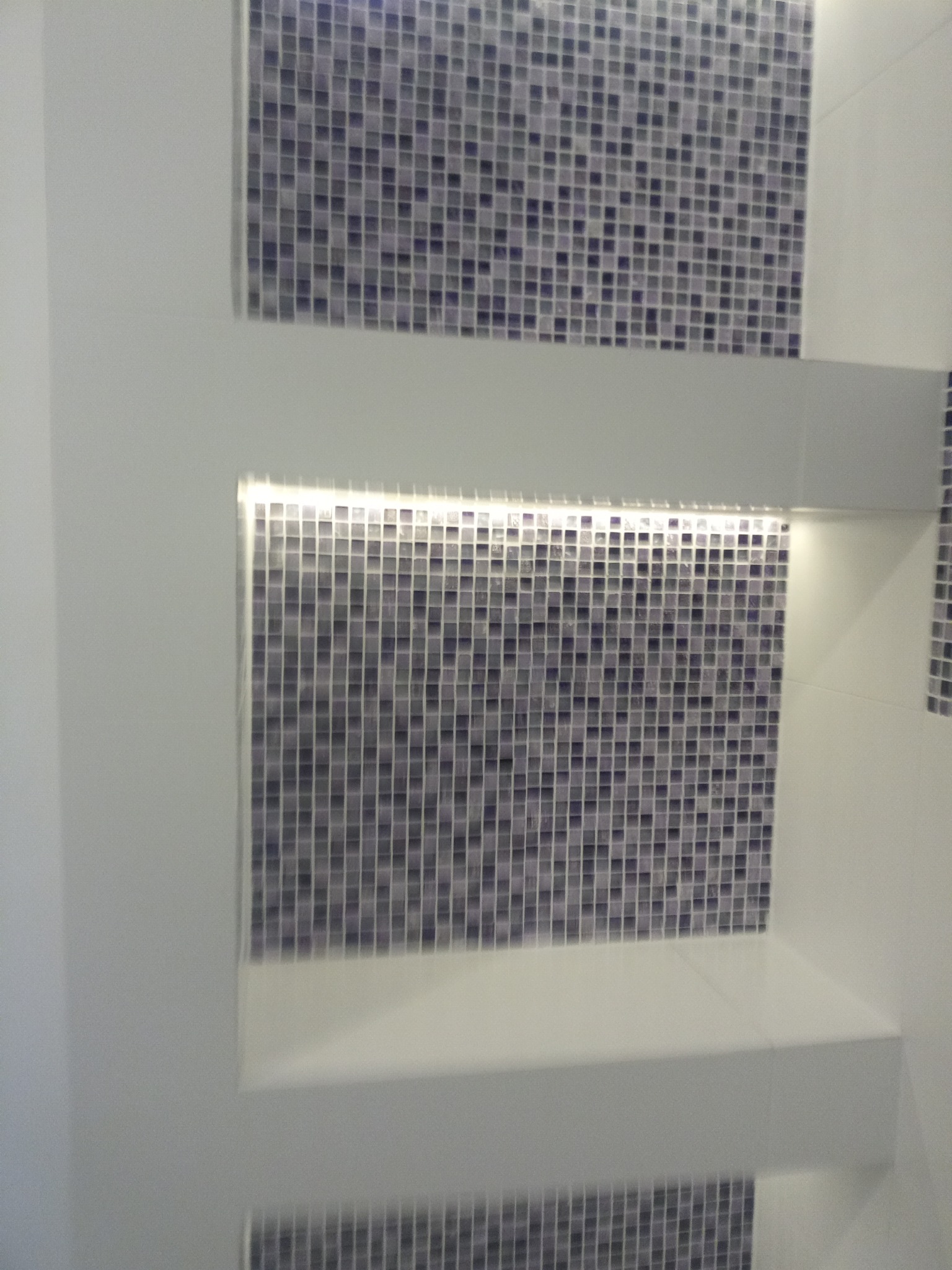 Fragment ściany łazienki z wnękami wyłożonymi mozaiką w odcieniach szarości i fioletu, oświetlonymi od góry punktowym światłem LED.