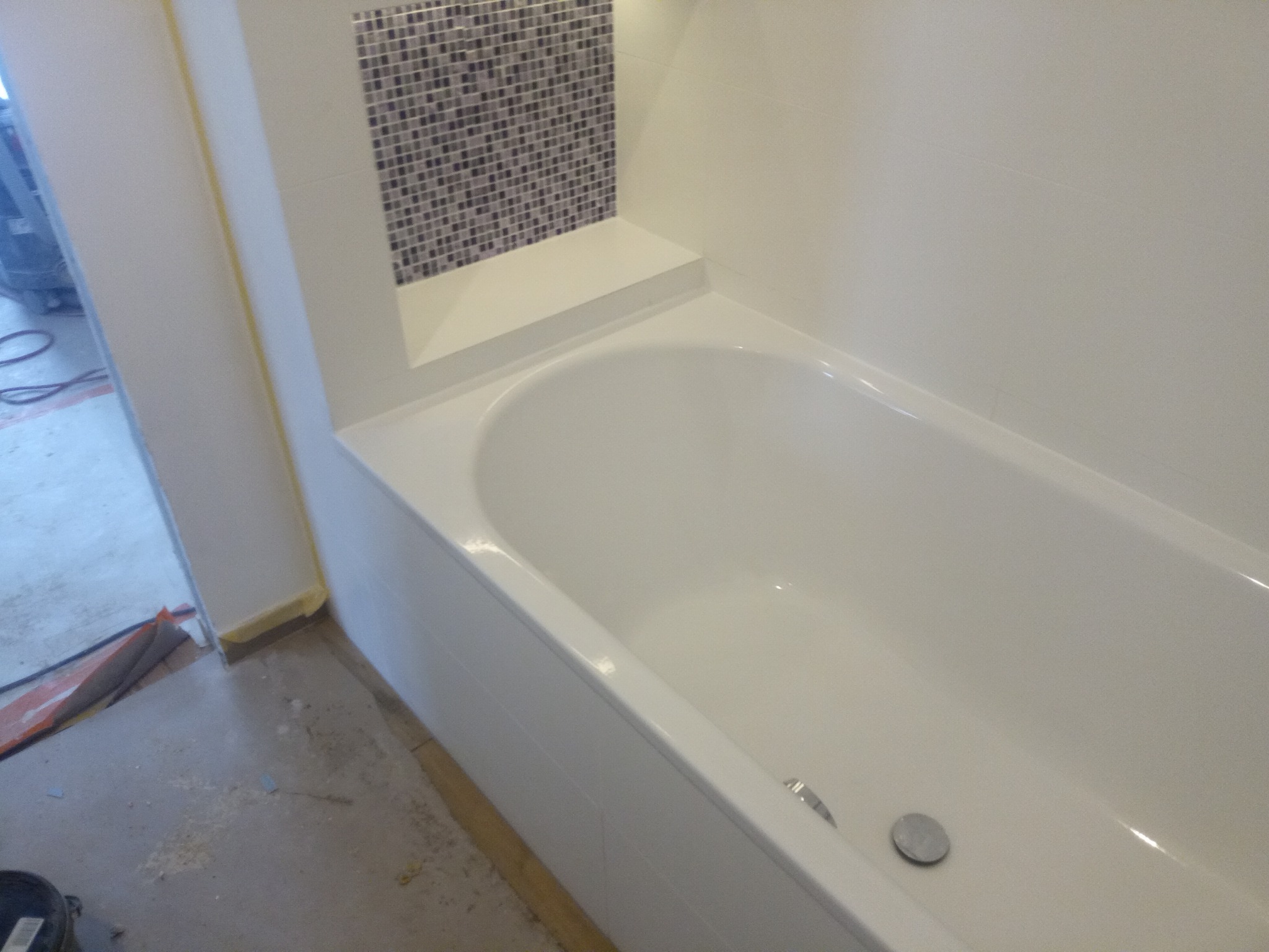 Biała wanna po renowacji, z mozaiką w strefie prysznicowej i półką, widoczne ślady prac wykończeniowych na podłodze.