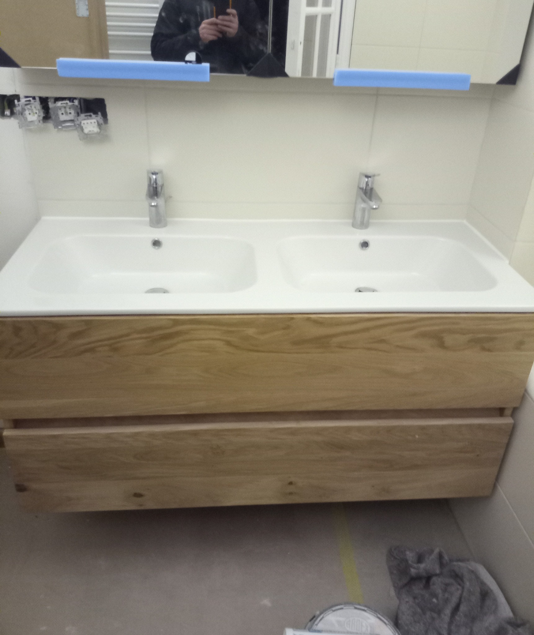 Nowoczesna umywalka podwójna z drewnianą szafką i chromowaną armaturą, widoczne niedokończone prace instalacyjne i odbicie osoby robiącej zdjęcie w lustrze.