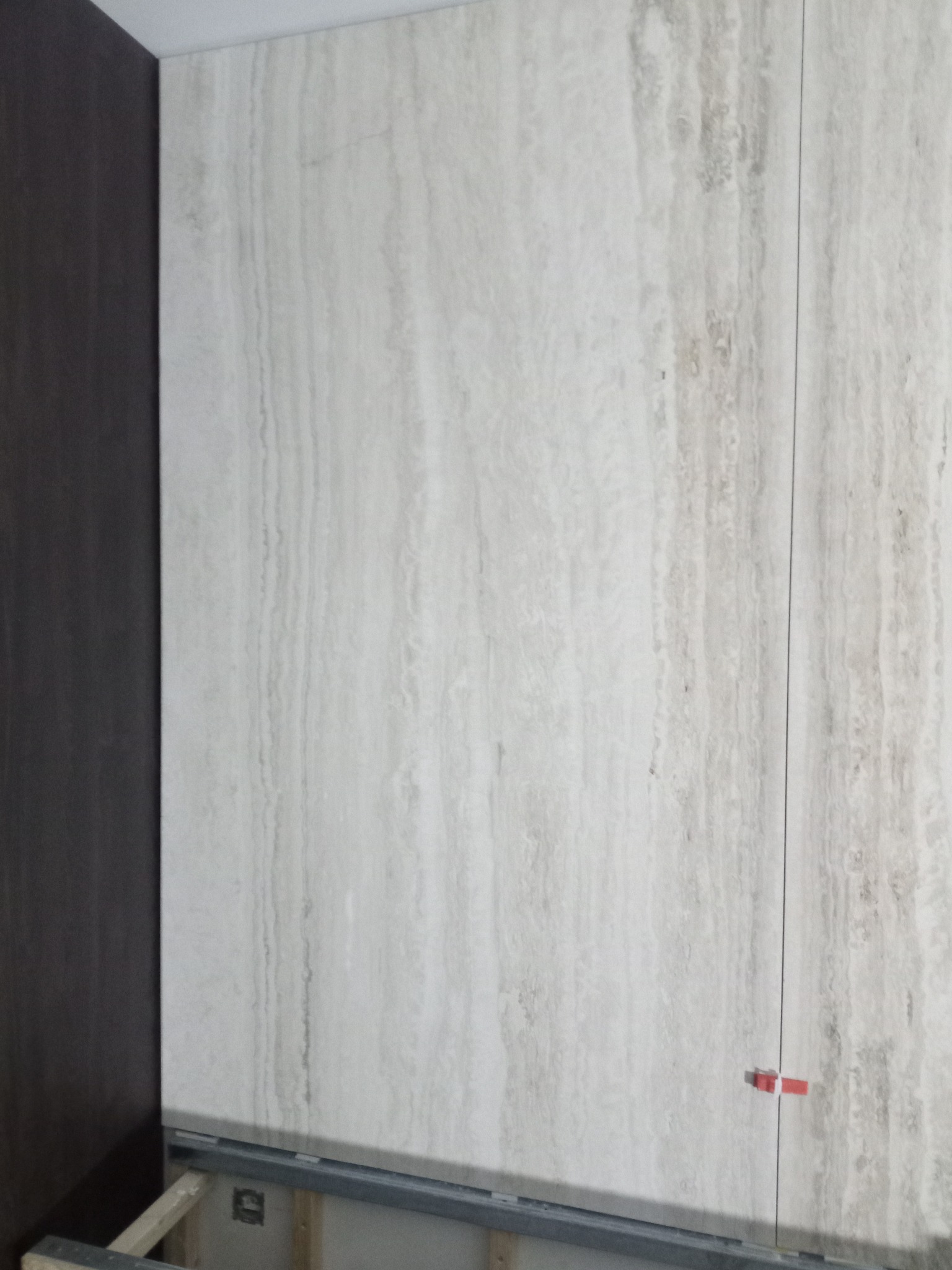 Ściana wyłożona płytami imitującymi trawertyn, z widocznymi elementami konstrukcji podwieszanej i czerwonymi krzyżykami dystansowymi między płytami.