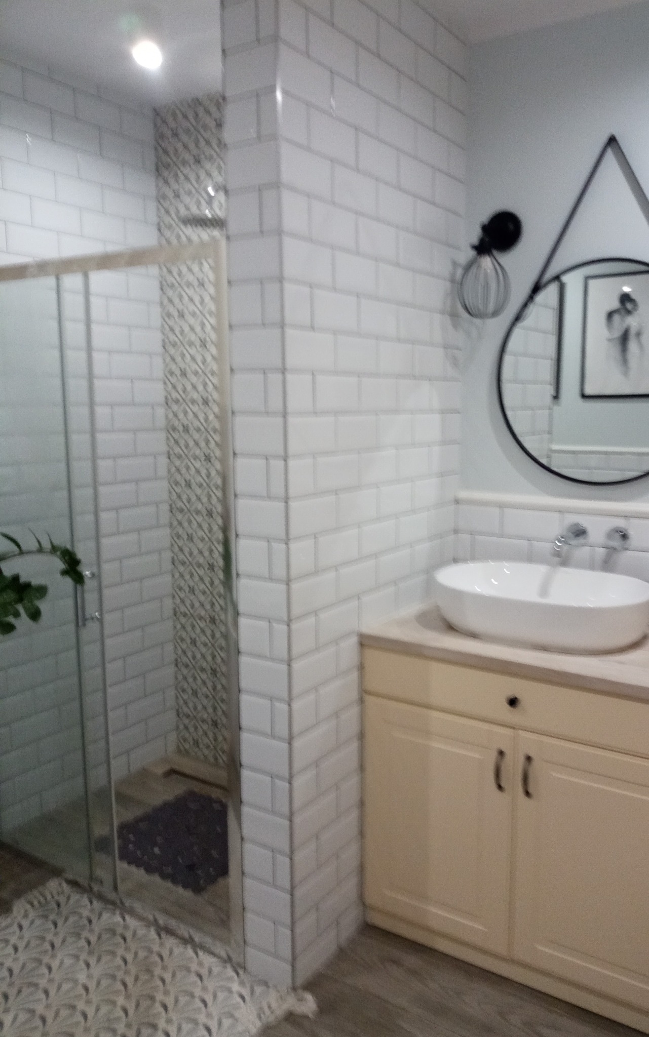 Wyremontowana łazienka z kabiną prysznicową wyłożoną białymi cegiełkami, umywalką nablatową na szafce w kolorze ecru, okrągłym lustrem w czarnej ramie i dekoracyjną lampą ścienną.
