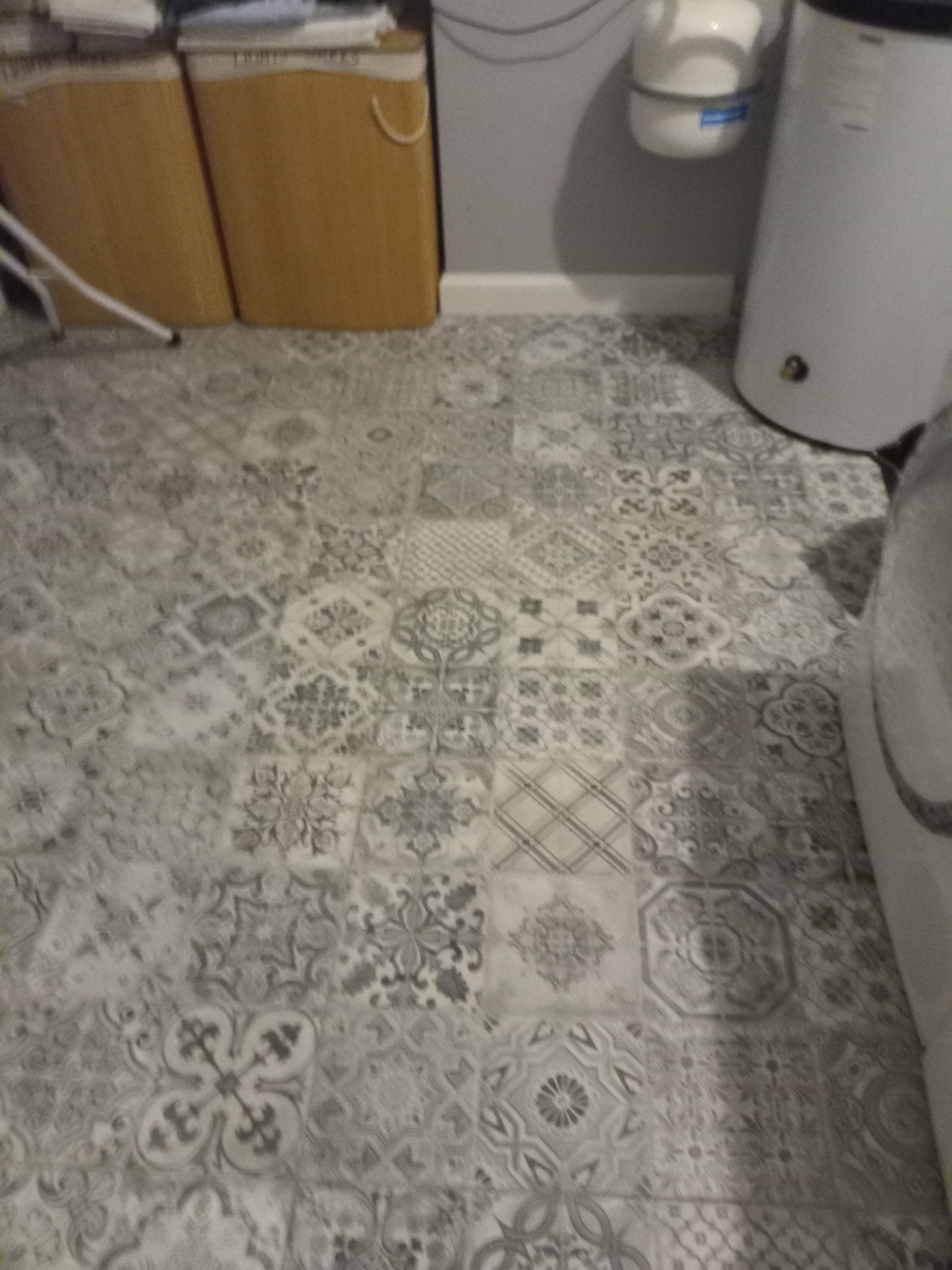 Szare płytki podłogowe z wzorem patchworkowym, widoczne kosze z praniem i bojler w tle.