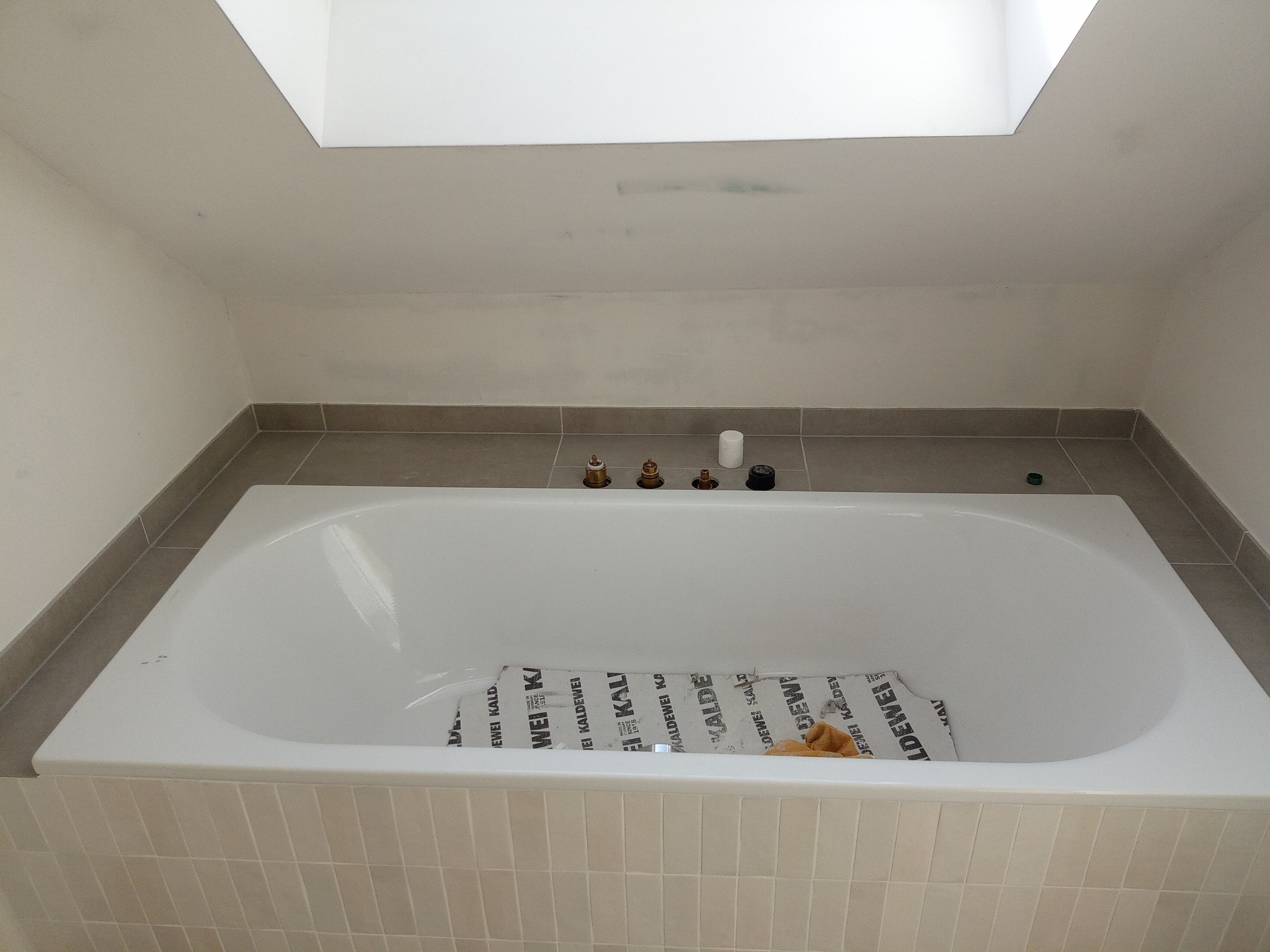 Nowa, biała wanna obudowana jasnymi płytkami, z zabezpieczeniem producenta wewnątrz, zamontowana w łazience z szarymi płytkami na ścianach i suficie z oknem dachowym.