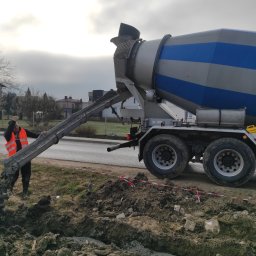 Przemysław Stachurski - Betoniarka w trakcie wylewania betonu na przygotowany teren. Pracownik w odblaskowej kamizelce nadzoruje proces. Widoczne wykopane rowy i ziemia.
