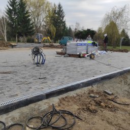 Przemysław Stachurski - Fragment placu z ułożoną szarą kostką brukową, widoczny drenaż liniowy, palety kostki i mini koparka w tle. Częściowo ukończony projekt z elementami wykończeniowymi.