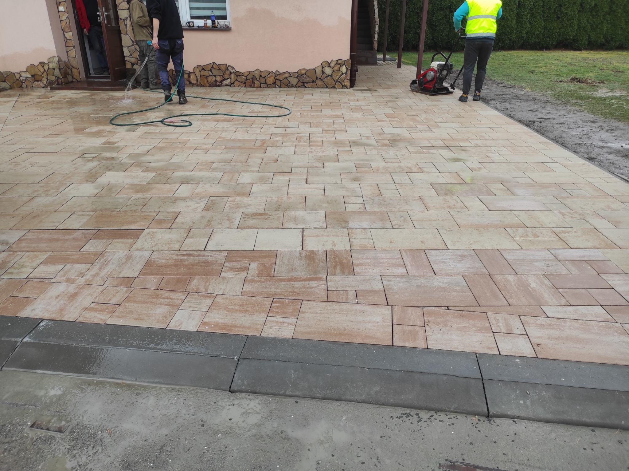 Nowa, beżowa kostka brukowa ułożona na podjeździe, częściowo mokra od wody z węża. W tle budynek z kamienną dekoracją i zagęszczarka.