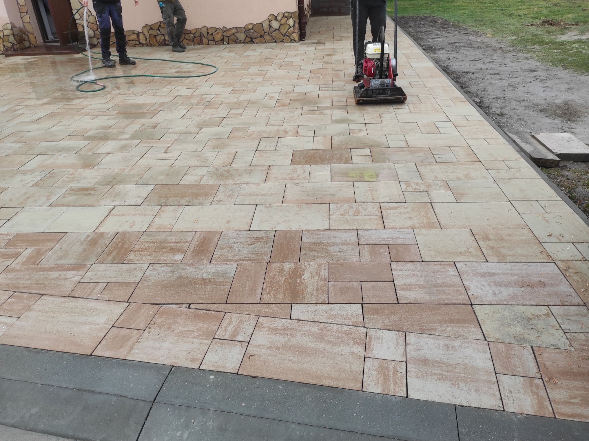 Układanka z kostki brukowej w odcieniach beżu i brązu, częściowo mokra od wody. W tle widoczna ubijarka wibracyjna i fragment budynku z kamienną elewacją.