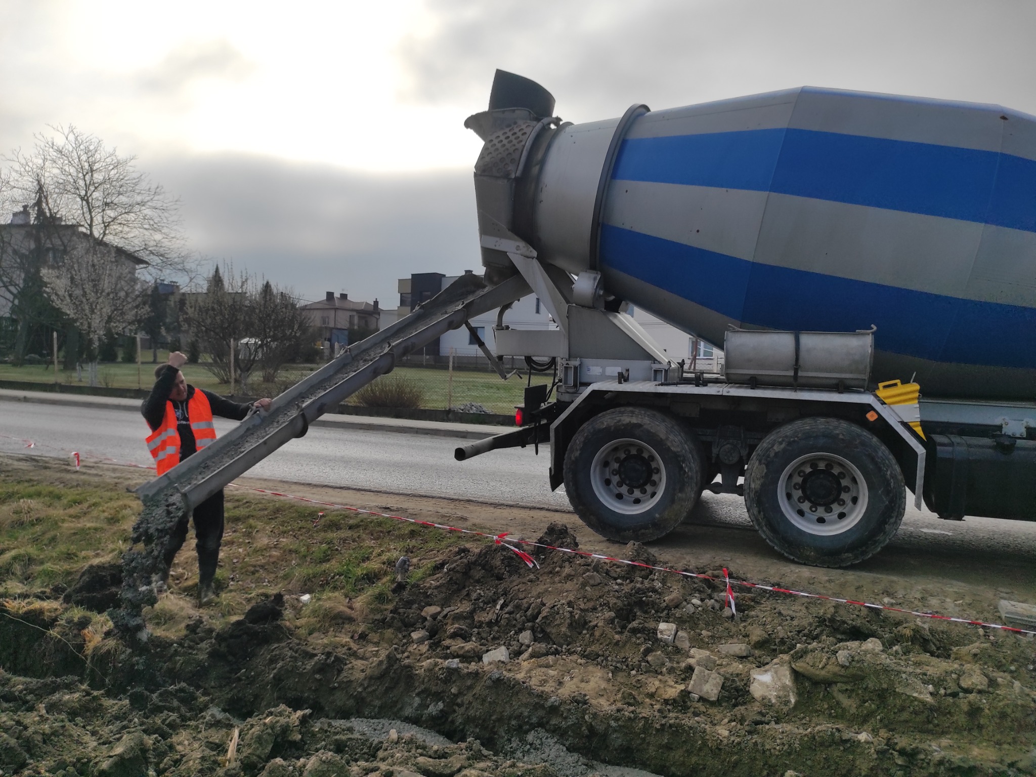 Betoniarka w trakcie wylewania betonu na przygotowany teren. Pracownik w odblaskowej kamizelce nadzoruje proces. Widoczne wykopane rowy i ziemia.