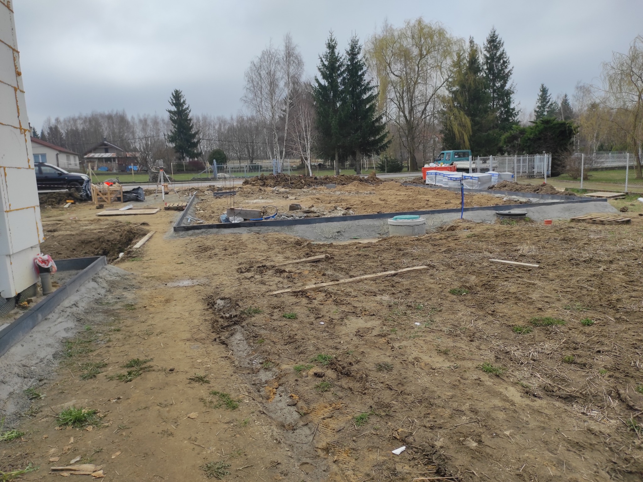 Przygotowanie terenu pod budowę: wytyczone fundamenty, widoczne obrzeża, zgromadzony materiał budowlany na działce. W tle drzewa i zabudowania.