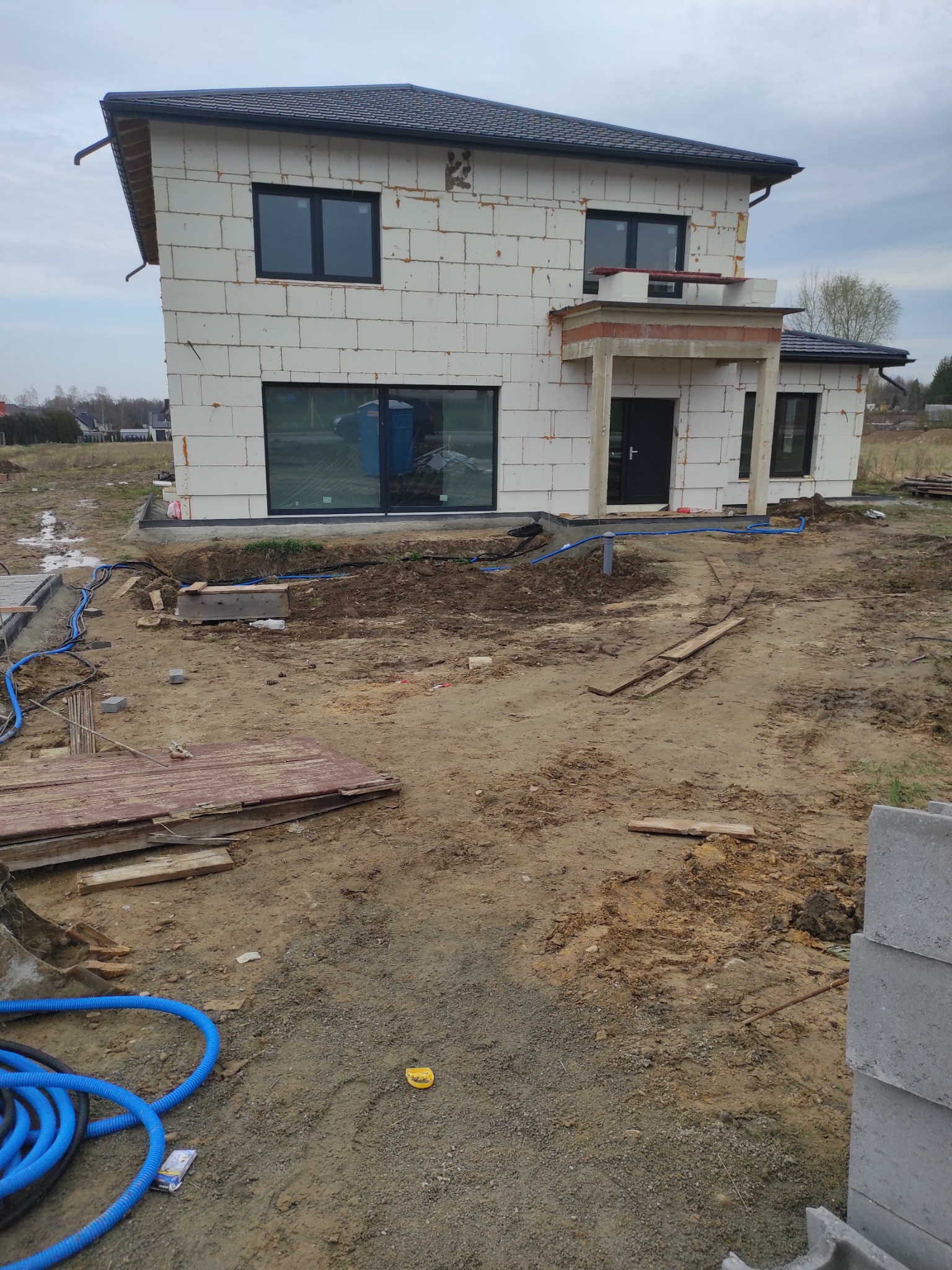Budowa domu z białego bloczka: widoczne fundamenty, instalacje w ziemi (niebieskie rury), częściowo wykończona elewacja, okna. Etap prac ziemnych i instalacyjnych.