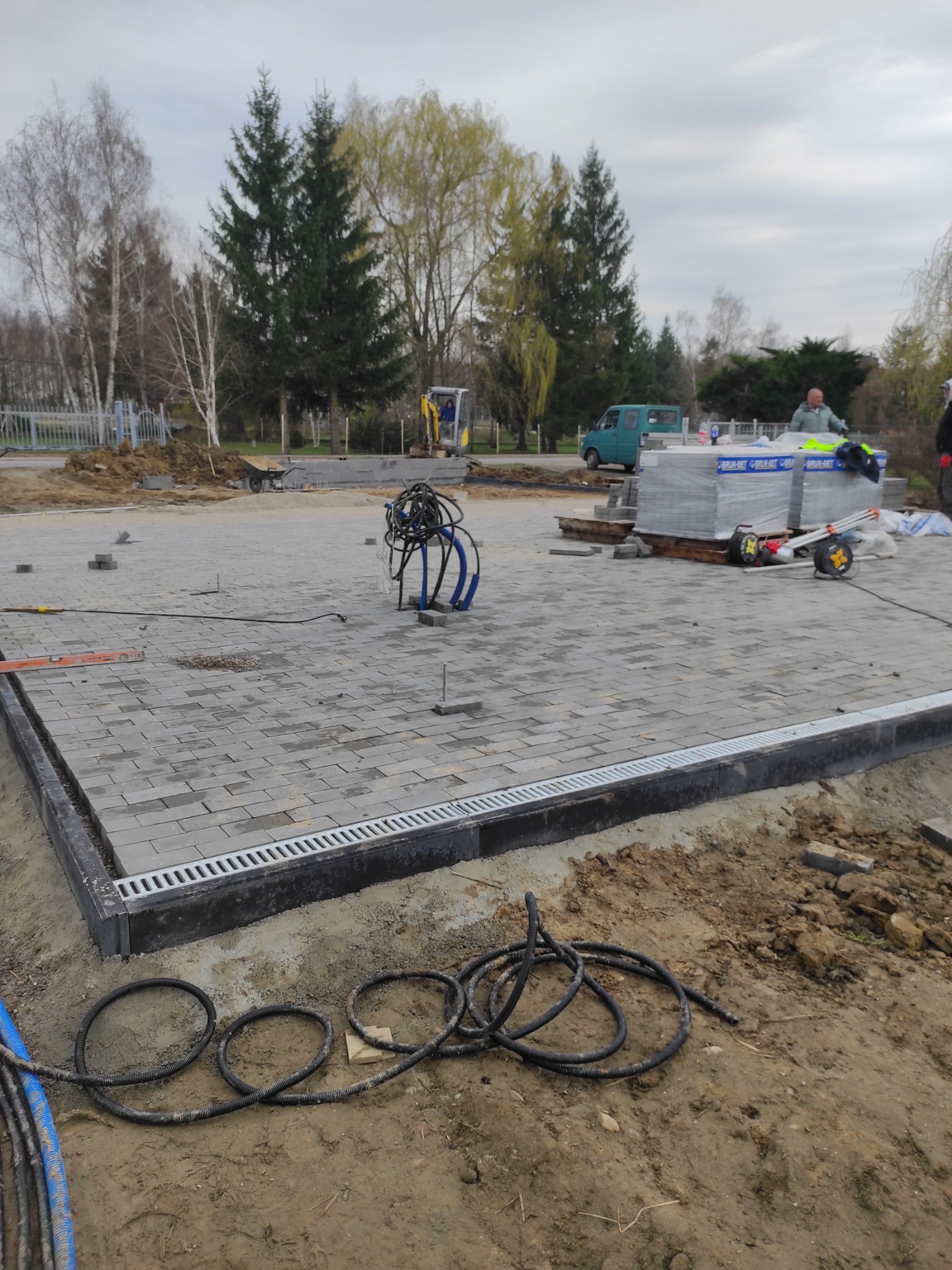 Częściowo ułożona szara kostka brukowa z odpływem liniowym. W tle widać materiały budowlane i mini koparkę, co sugeruje trwające prace.