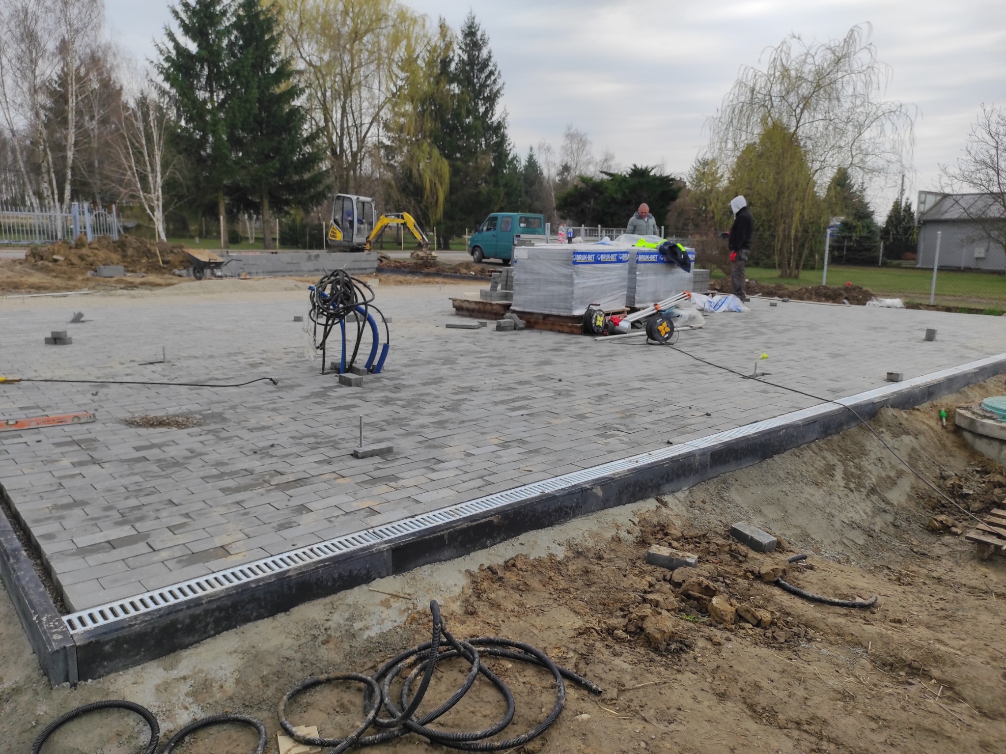 Fragment placu z ułożoną szarą kostką brukową, widoczny drenaż liniowy, palety kostki i mini koparka w tle. Częściowo ukończony projekt z elementami wykończeniowymi.