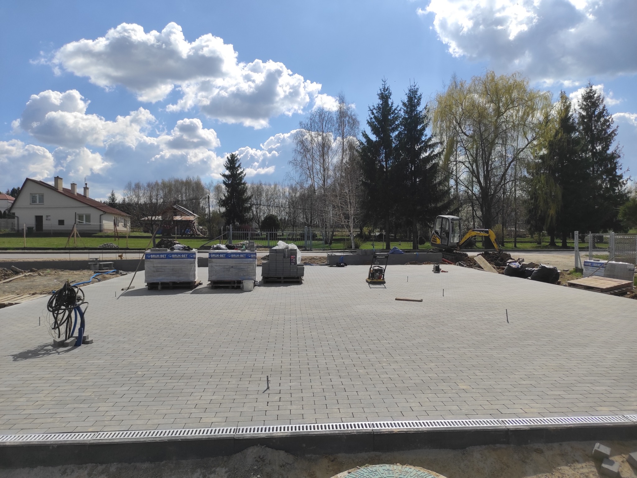 Ułożona szara kostka brukowa na placu budowy w słoneczny dzień. Palety kostki, drenaż liniowy, minikoparka w tle. Widok z góry na ukończony etap prac.