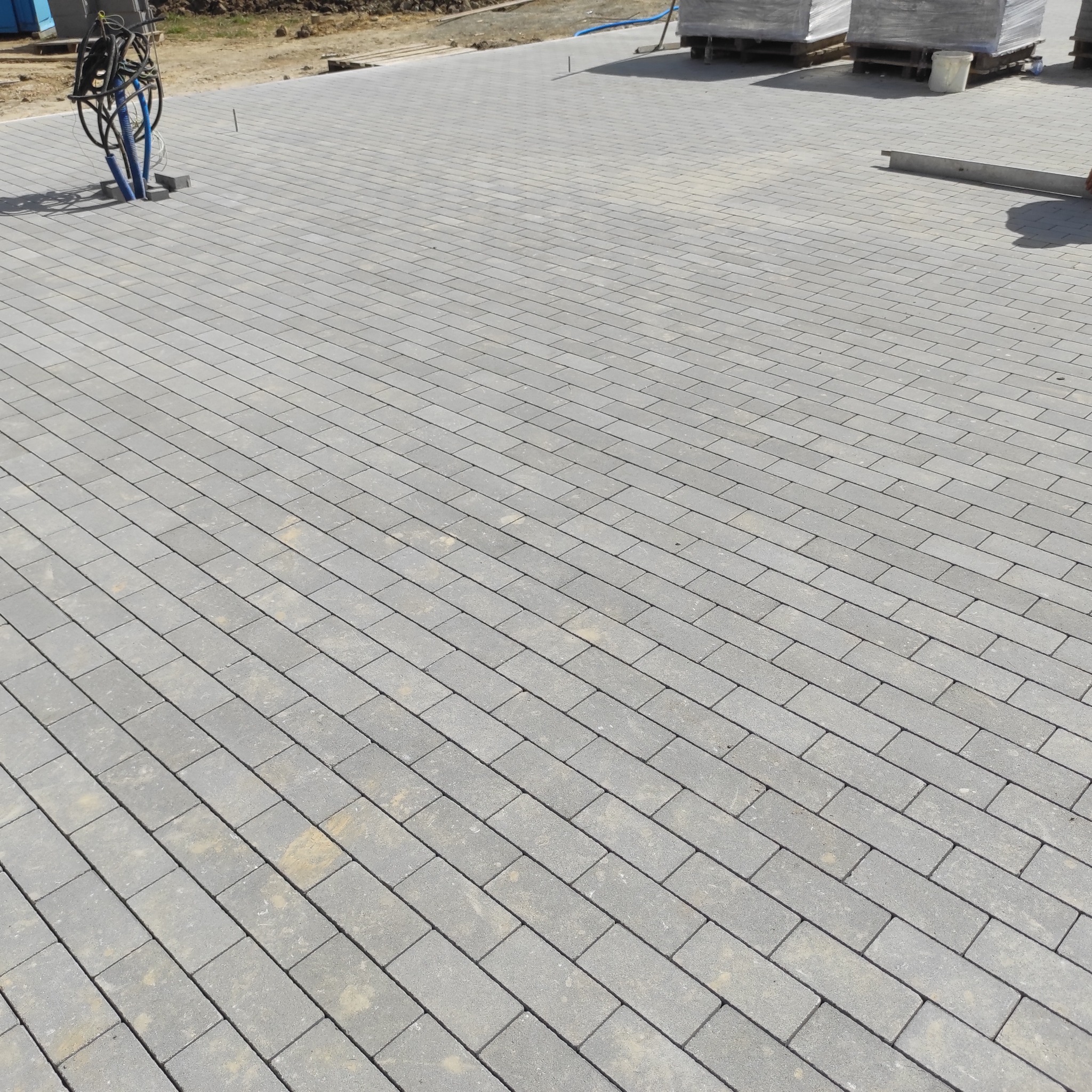 Świeżo ułożona, szara kostka brukowa w prostokątnym wzorze. Widoczne palety z kostką i wystające rury instalacyjne. Jasne, naturalne światło.
