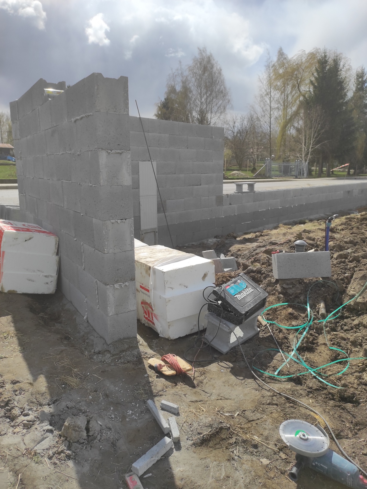 Wznoszenie muru z bloczków betonowych na fundamencie. Widoczne narzędzia, materiały izolacyjne i fragment otoczenia z zielenią. Budowa w toku, etap murowania.