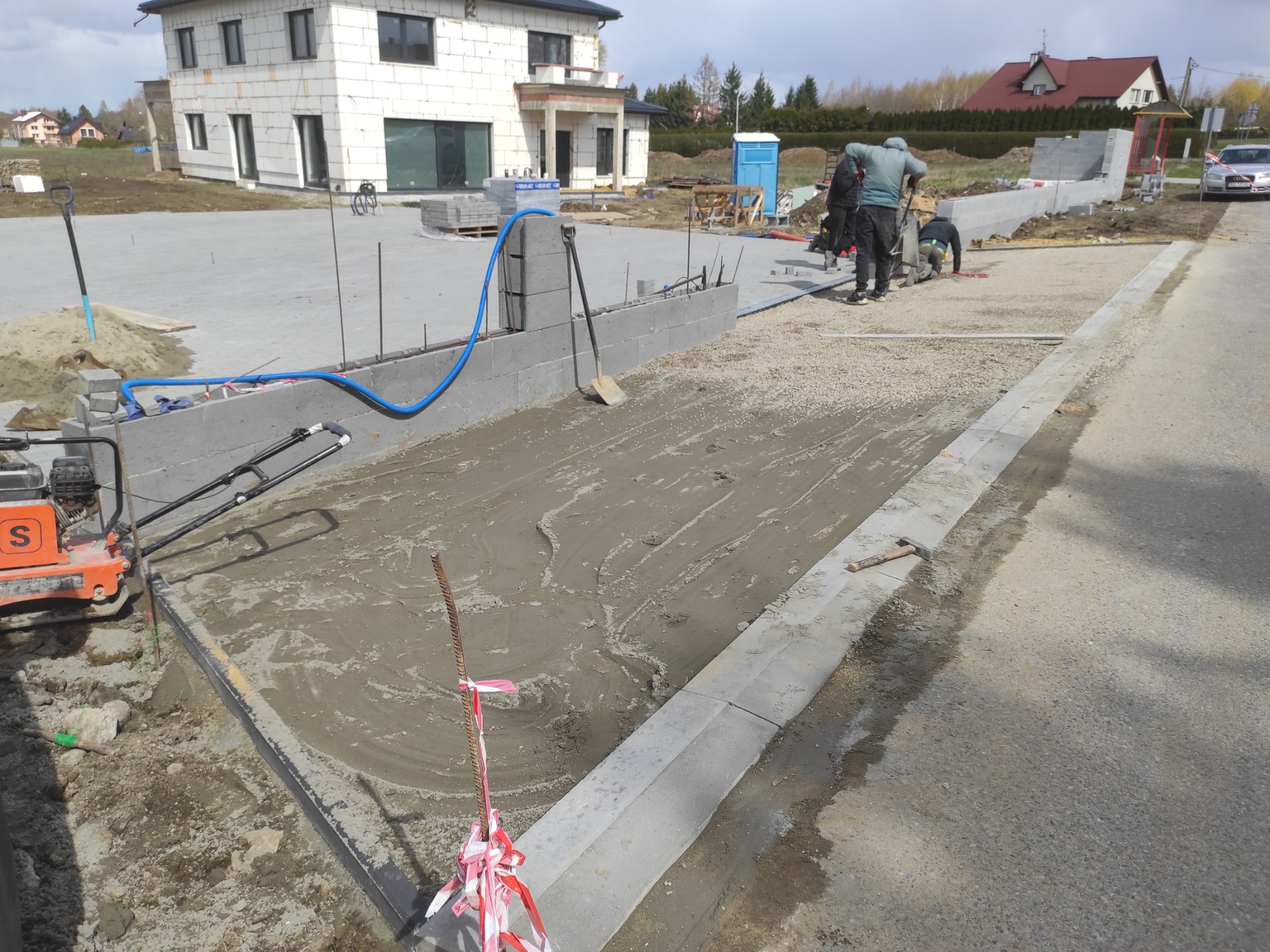 Przygotowanie podłoża pod chodnik z kostki: ubijanie piasku zagęszczarką, obrzeża chodnikowe, w tle budynek w stanie surowym otwartym i ekipa pracowników.