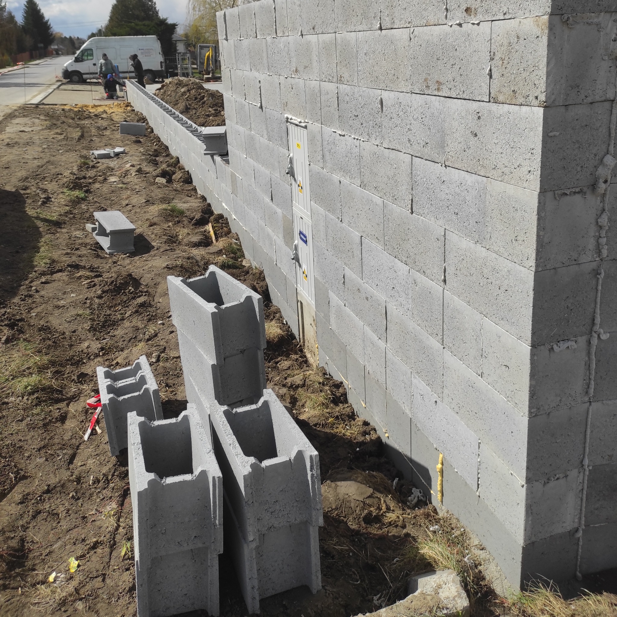 Wznoszenie ściany z szarych bloczków betonowych. Widoczne zapasy materiału i ekipa budowlana w tle. Częściowo ukończona konstrukcja z widocznymi spoinami.